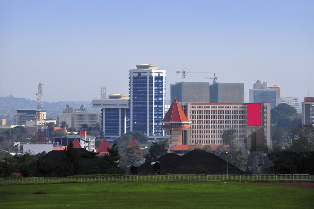 City skyline with blue glass towers behind a grassy foreground and red-roofed buildings in front, with construction cranes in the distance.